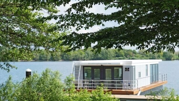 Une péniche moderne dotée d’une terrasse et de fenêtres, entourée de verdure et d’eau.