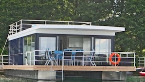 A floating houseboat with a deck, seating area, and a lifebuoy.