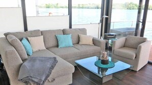 A modern living room with a sofa and armchair, a glass coffee table, and a view of a body of water through large windows.