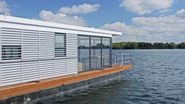 A modern floating house with a wooden deck and glass windows overlooking a lake. - Hausboot am Xantener Nordsee (Xanten)