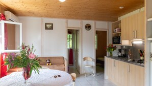 Ein Zimmer mit Holzbalkendecke, einem Esstisch mit Blumenvase, einer Couch, einer Küchenzeile inklusive Schränken und Geräten sowie einem roten Vorhang.