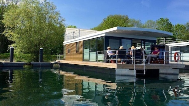 A modern boat with a covered deck and glass walls, surrounded by calm water and greenery.