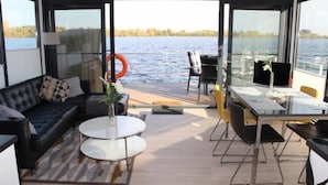 A modern living area with a sofa, two round tables, and a dining table with chairs. A large window offers a view of a body of water and trees.