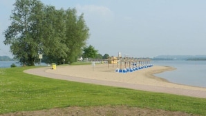 A lakeside park with a sandy path, blue seating, and a large tree.
