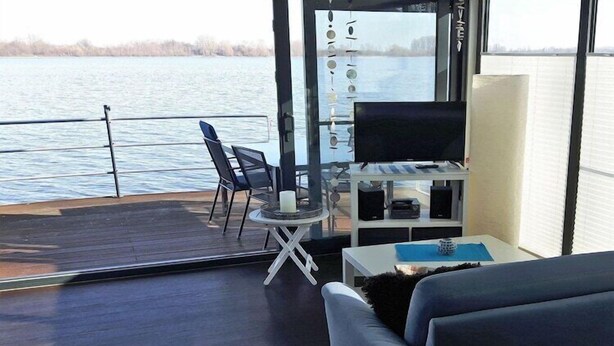 A modern living area with a sofa, a small table, and a TV on a balcony overlooking a body of water.