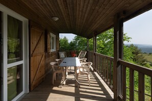 Um deck de madeira com mesa e cadeiras, janelas amplas e vista para o verde.