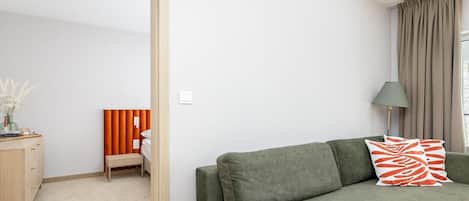 A modern living room with a green sofa, a bench, a lamp, and a view of a bedroom with a bed and curtains.