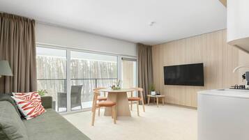 A modern living room with a sofa, a dining table with chairs, a flat-screen TV mounted on the wall, and a large window with curtains.