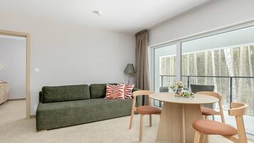 A modern living room with a green sofa, a round wooden table with chairs, and a large window offering a view of trees.