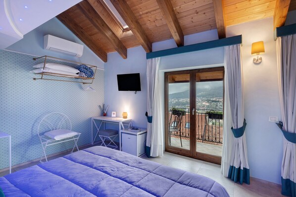 A bedroom with a bed, a desk, a chair, a TV, and a balcony with curtains.