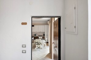 Uma porta dá acesso a uma cozinha com armários brancos, uma mesa de jantar com cadeiras e um aviso na parede.