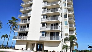 A tall, multi-story hotel with balconies, a paved entrance, and palm trees in the background.