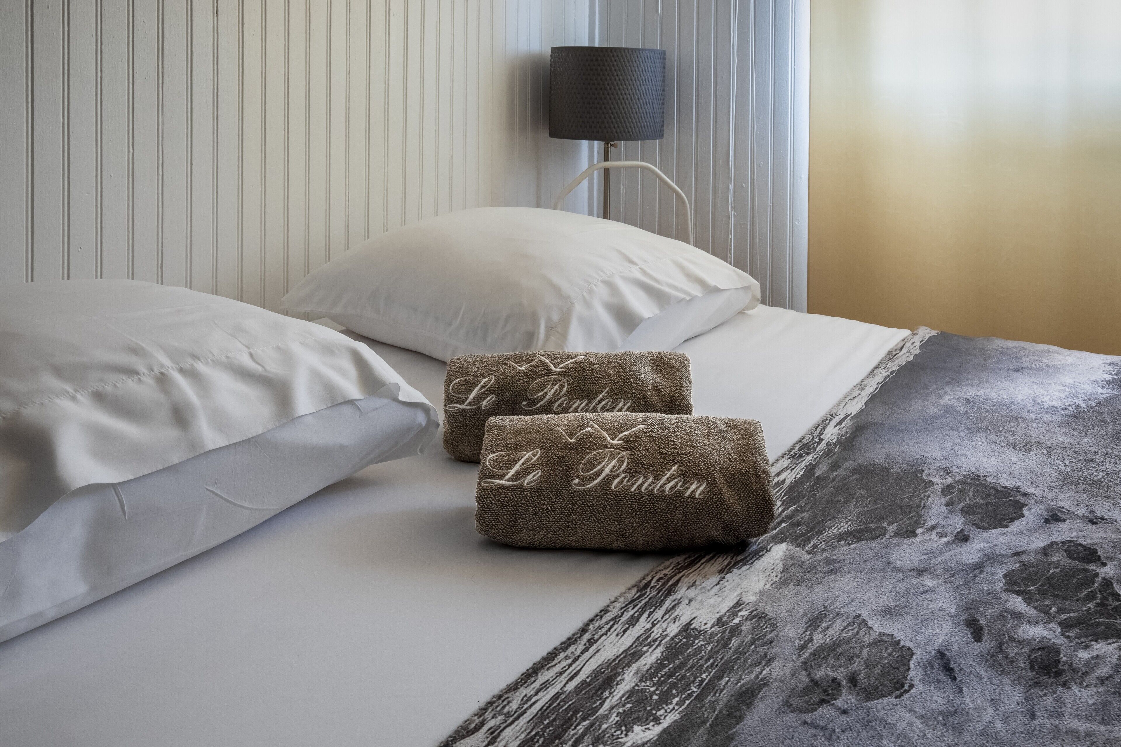A bed with white linens, two brown towels with 'Le Ponton' text, a lamp, and a striped wall.