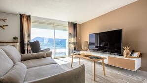 A modern living room with a large TV, a sofa, a coffee table, and a view of the sea.