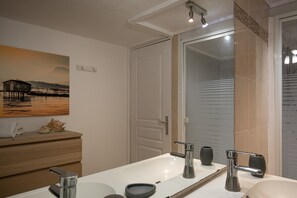 Une salle de bain avec un grand miroir, un lavabo blanc, un espace douche et un meuble-lavabo en bois sur lequel est posée une serviette.