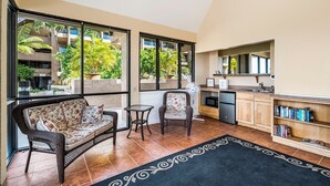 Amplia sala de estar con sofá, sillón y una mesita, ventanas grandes con vistas a un edificio y zonas verdes, y un área de cocina con fregadero y estanterías.