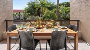 Una mesa de comedor de madera preparada para cuatro personas, con sillas de mimbre, rodeada por una barandilla de cristal, con vistas a un jardín con palmeras y un edificio al fondo.
