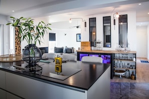 A modern kitchen with a central island, a dining area with a sofa and chairs, and a bar area with shelves and glassware. - Exceptional Villa – Les Pieds dans l’Eau in Saint-Mandrier-sur-Mer (Saint-Mandrier-sur-Mer)