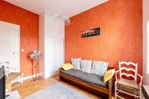 Une pièce comprenant un lit, une chaise, un bureau, un ventilateur sur pied et un tableau accroché au mur.