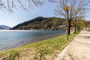 Uma trilha à beira do lago com um lampião, gramado e um morro com edifícios.