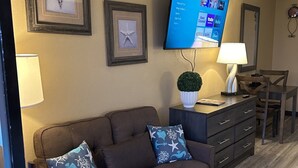 A living room with a brown sofa, two decorative pillows, a flat-screen TV mounted on the wall, a small potted plant, a lamp, and a wooden dining table with chairs. - Stylish ocean view 2 queen efficiency condo in marvelous Myrtle Beach (Myrtle Beach)