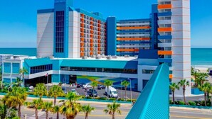 A modern hotel with a prominent blue and orange facade, multiple floors, and a parking area with palm trees. - Stylish ocean view 2 queen efficiency condo in marvelous Myrtle Beach (Myrtle Beach)