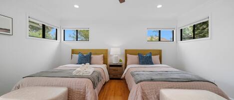 A hotel room with two beds, a ceiling fan, a nightstand with a lamp, and two windows with views of greenery.