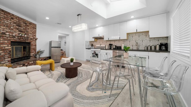 A modern living room with a brick fireplace, a sofa, and a glass dining table with chairs. - @ Marbella Lane - Serene and Pristine Home (West Covina)