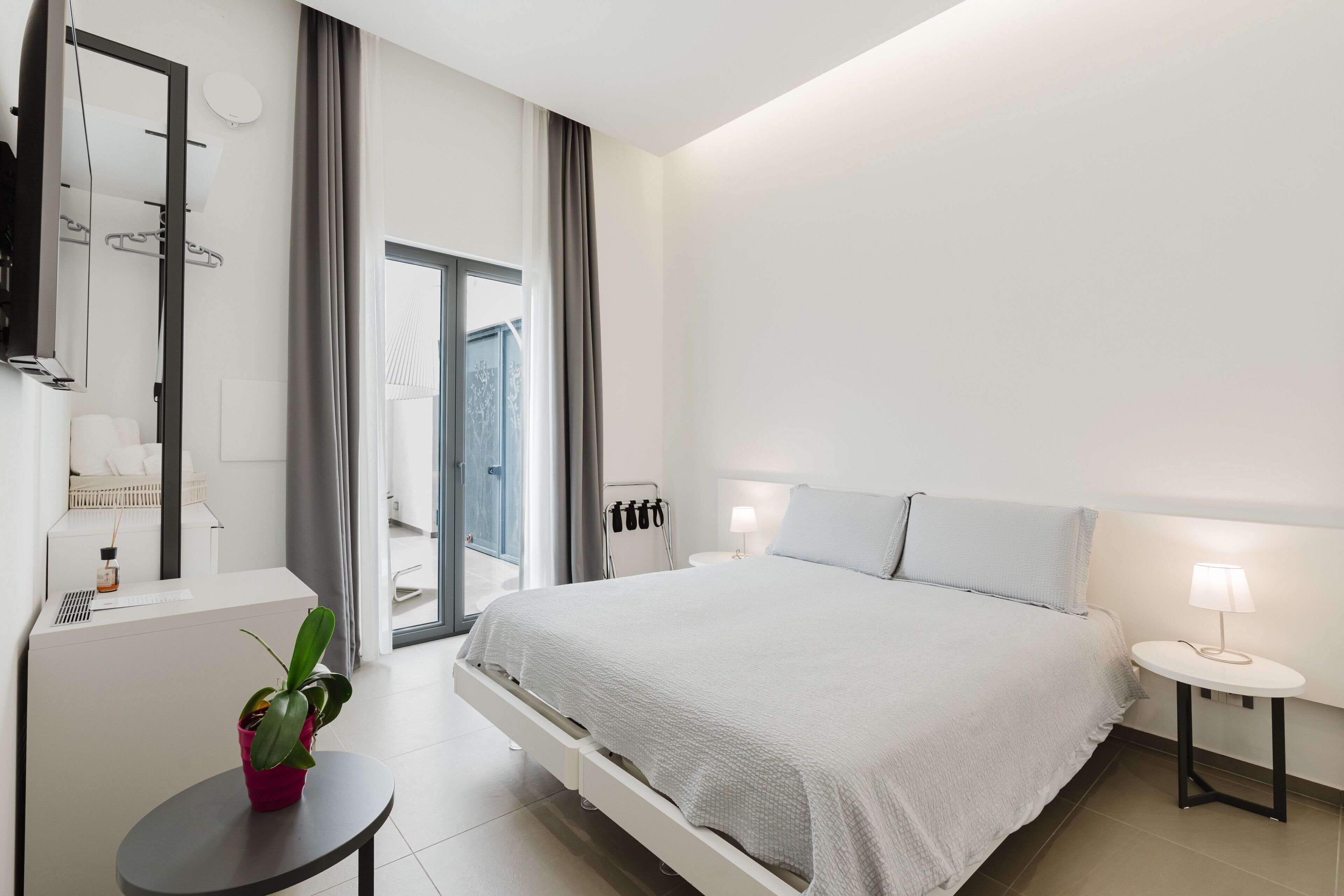 A modern hotel room with a bed, bedside table, lamp, potted plant, and a large window.
