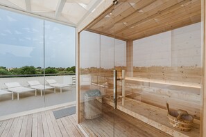 A modern wooden sauna with glass walls, a wooden deck, and white lounge chairs.