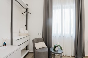 Une chambre d’hôtel moderne avec un fauteuil gris, une petite table ronde ornée d’une plante, une commode blanche et un miroir à cadre noir.