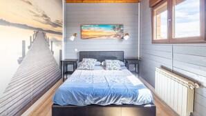 Un dormitorio con techo de madera, una cama con colcha azul, un cuadro de un atardecer y una ventana con vistas.