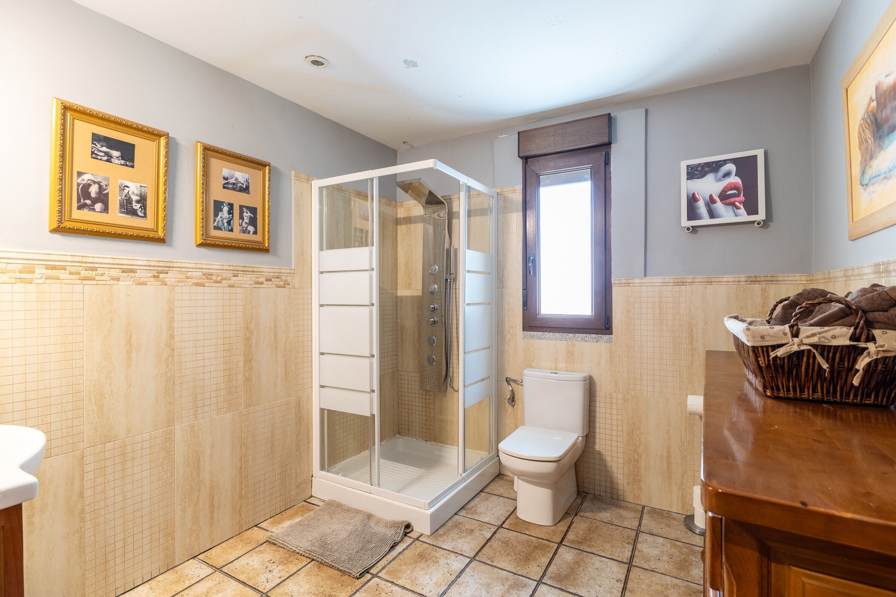 Un baño con ducha acristalada, inodoro y lavamanos. Hay fotografías enmarcadas en la pared.