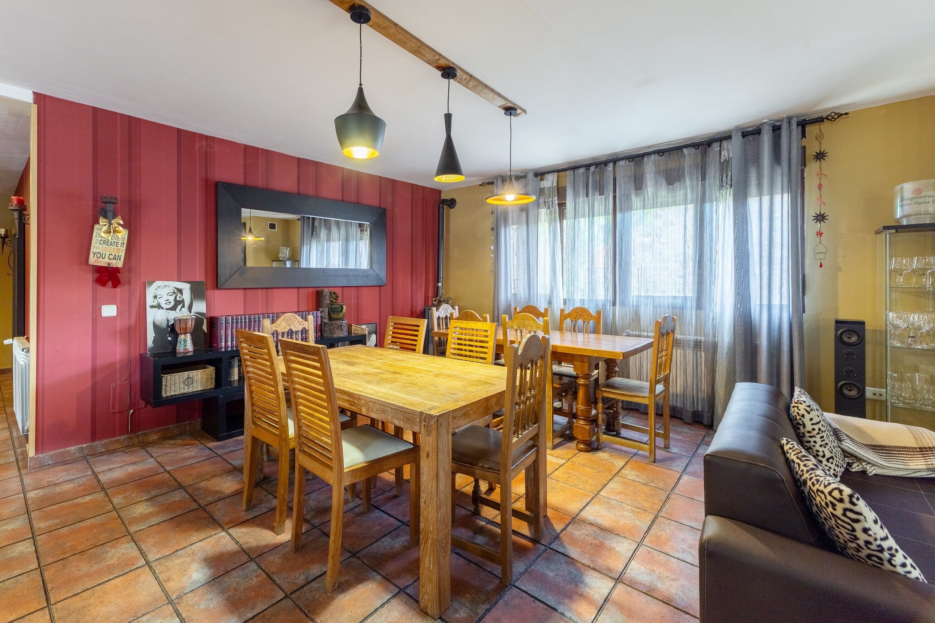 A dining area with wooden tables and chairs, a mirror on the wall, and a sofa with patterned cushions.