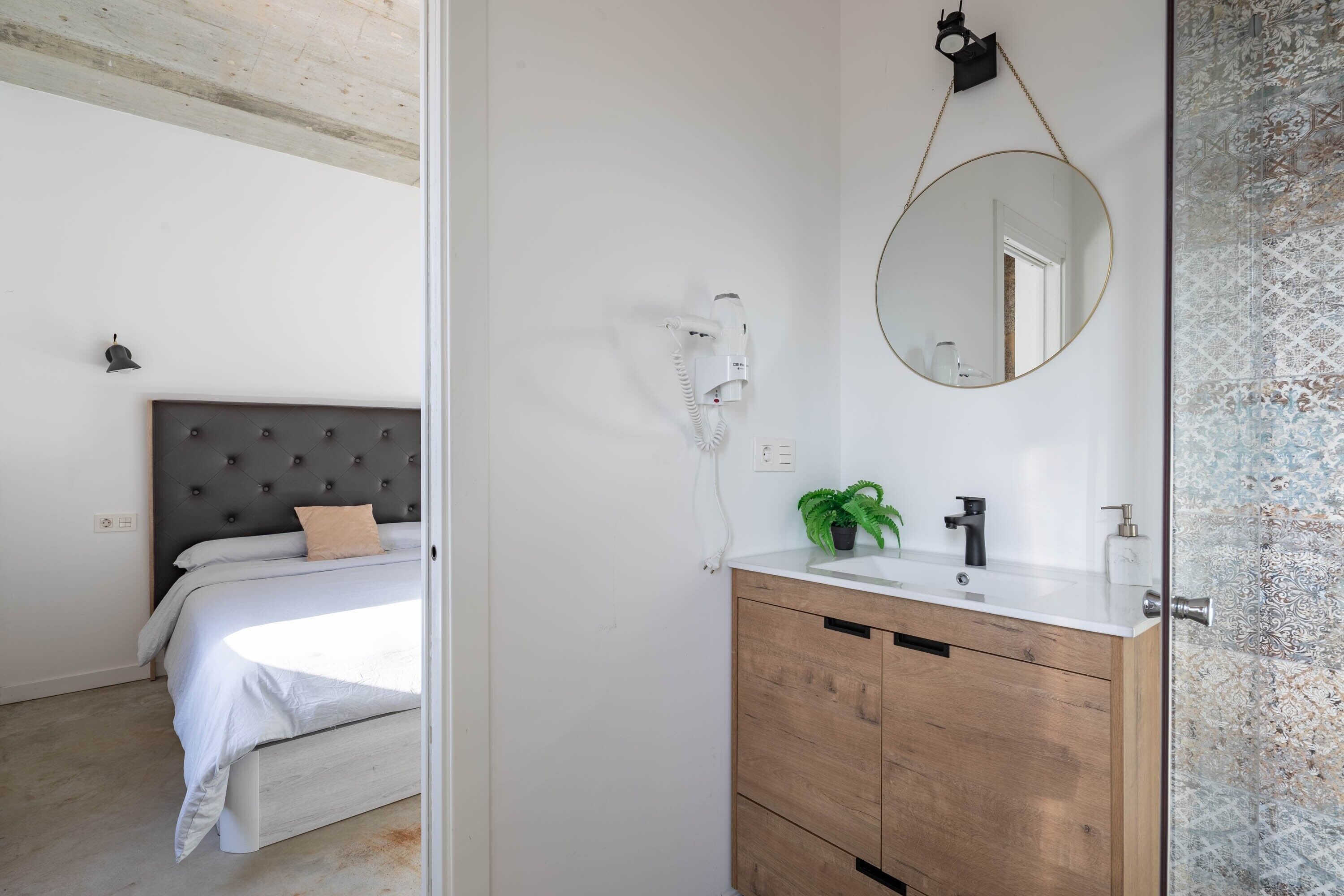 A bedroom with a bed, a bedside table with a lamp, and a bathroom with a sink, mirror, and towel rack.