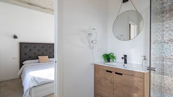 A bedroom with a bed, a bedside table with a lamp, and a bathroom with a sink, mirror, and towel rack.