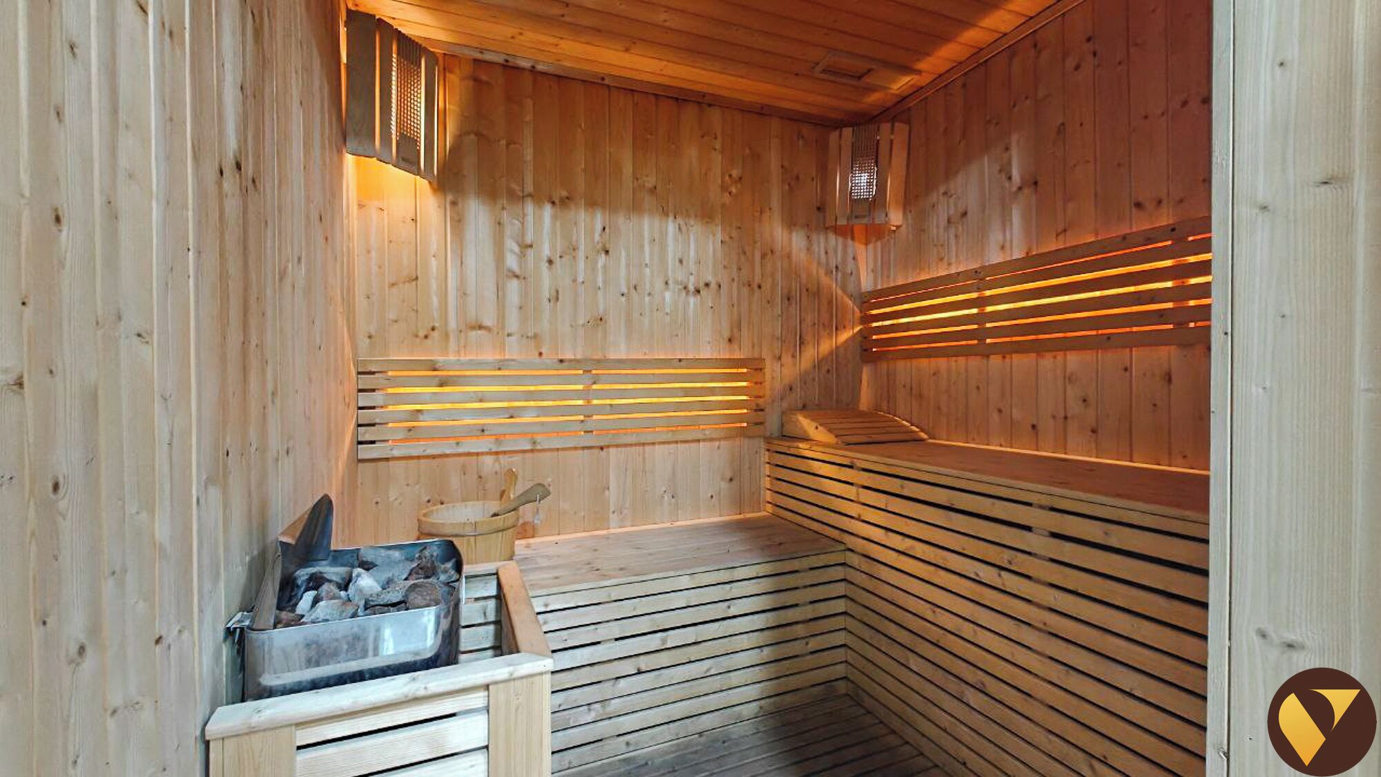 A sauna with wooden walls and benches, a bucket of stones, and a wooden bench with a metal bucket.