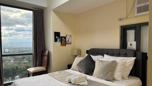 A hotel room with a large bed, a chair, a desk, and a view of the city. - 1BR Condo w/ Great View Near Venice Canal & BGC  (Taguig)