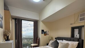 A hotel room with a bed, a desk, a chair, a window with curtains, and a view of the city. - 1BR Condo w/ Great View Near Venice Canal & BGC  (Taguig)
