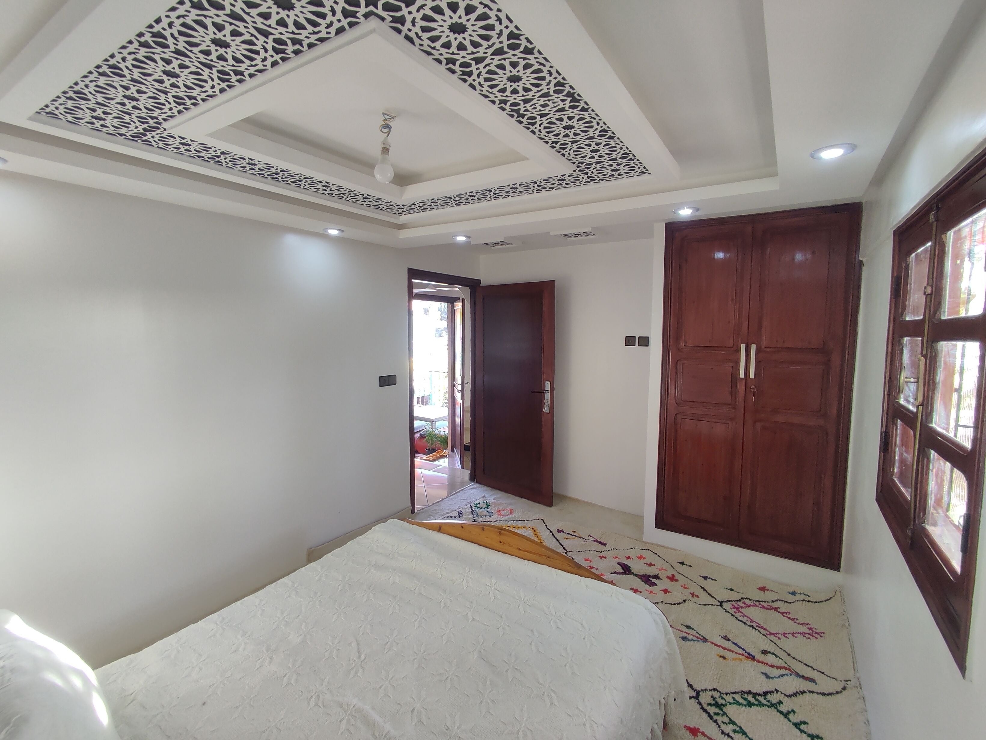 A room with a patterned ceiling, a bed with a white bedspread, a colorful rug, and wooden doors.