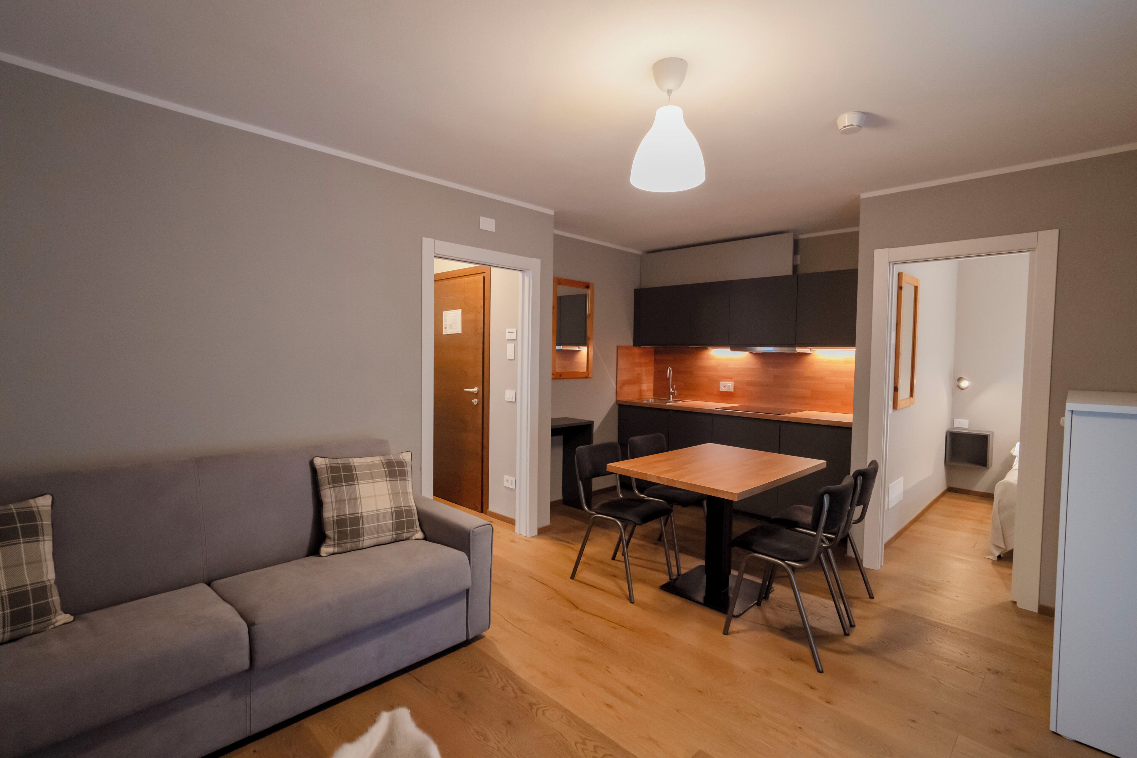 A modern living room with a sofa and a dining table, a kitchen area with wooden cabinets, and a small refrigerator.