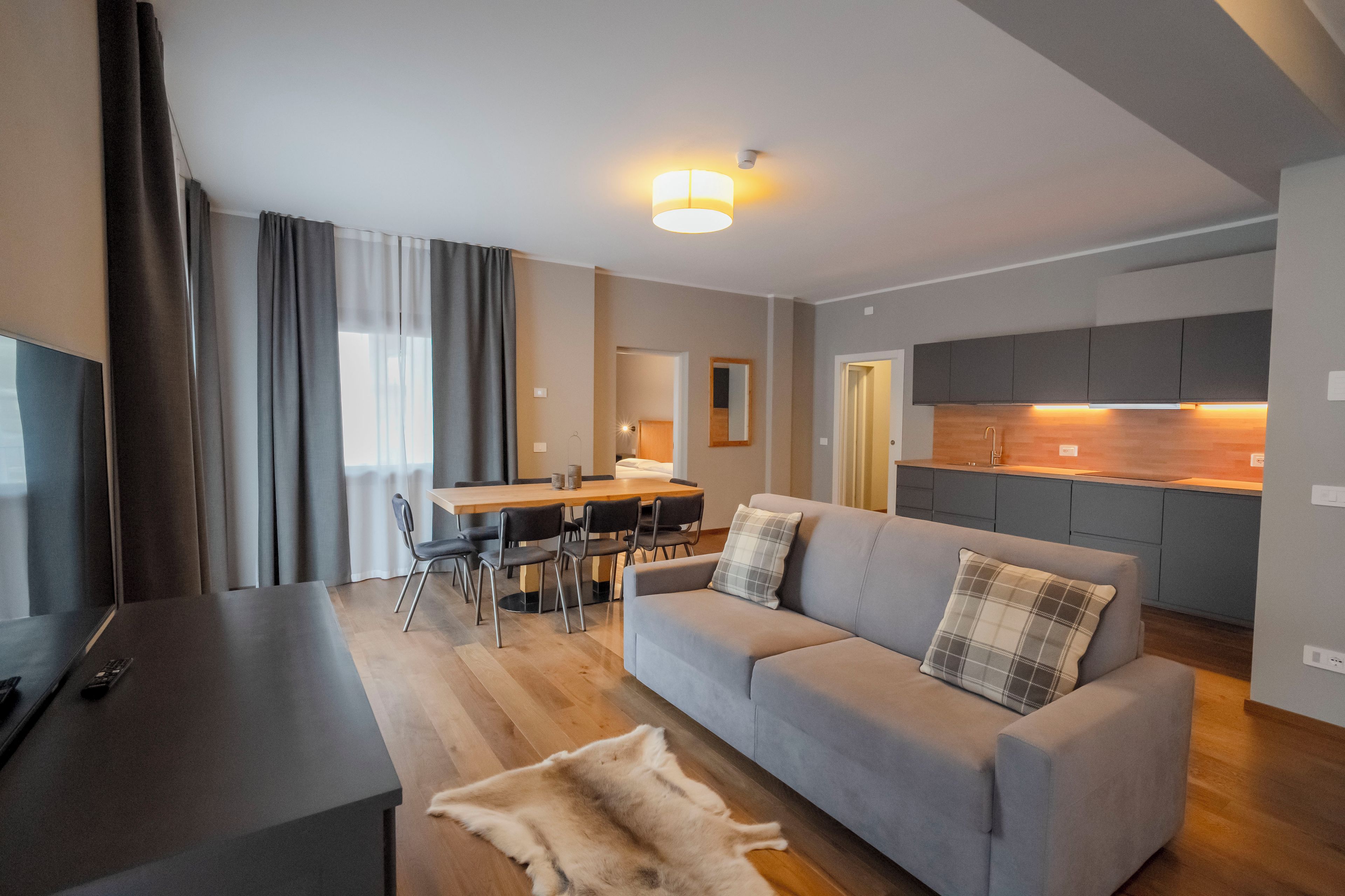 A modern living room with a grey sofa, a flat-screen TV, and a dining area with a wooden table and chairs.