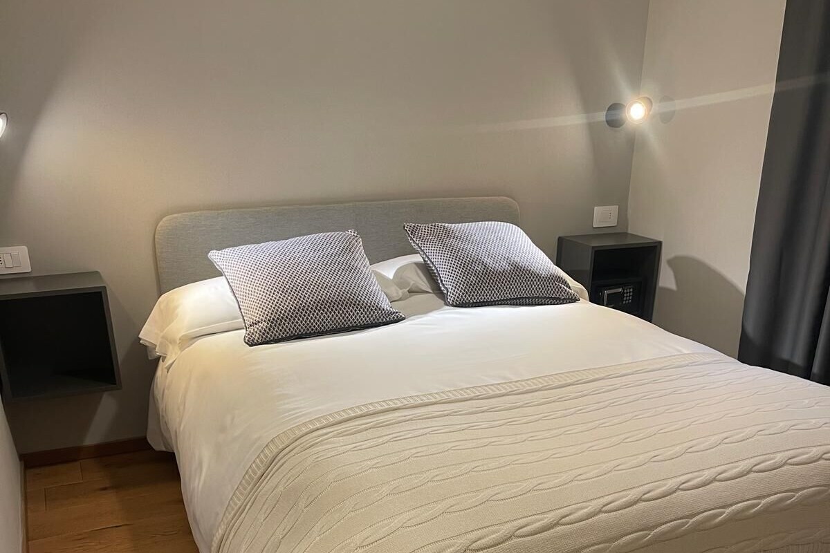 A neatly made bed with a white duvet and two patterned pillows, a wooden floor, a white furry rug, and a nightstand with a lamp.