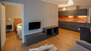 A modern living room with a flat-screen TV, built-in shelves, a sofa, and a kitchen area with grey cabinets and a wooden countertop. - Condhotel Fanti Ski & Bike (Valdisotto)