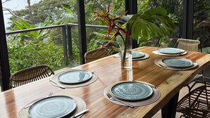 Une table à manger en bois, dressée avec des assiettes et des couverts, entourée de chaises en osier, avec un luminaire suspendu au-dessus et une vue sur une végétation luxuriante à l’extérieur.