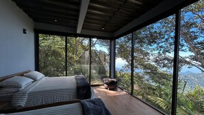 Un dormitorio moderno con dos camas, piso de madera y ventanas amplias que ofrecen vistas a vegetación exuberante y un paisaje lejano.