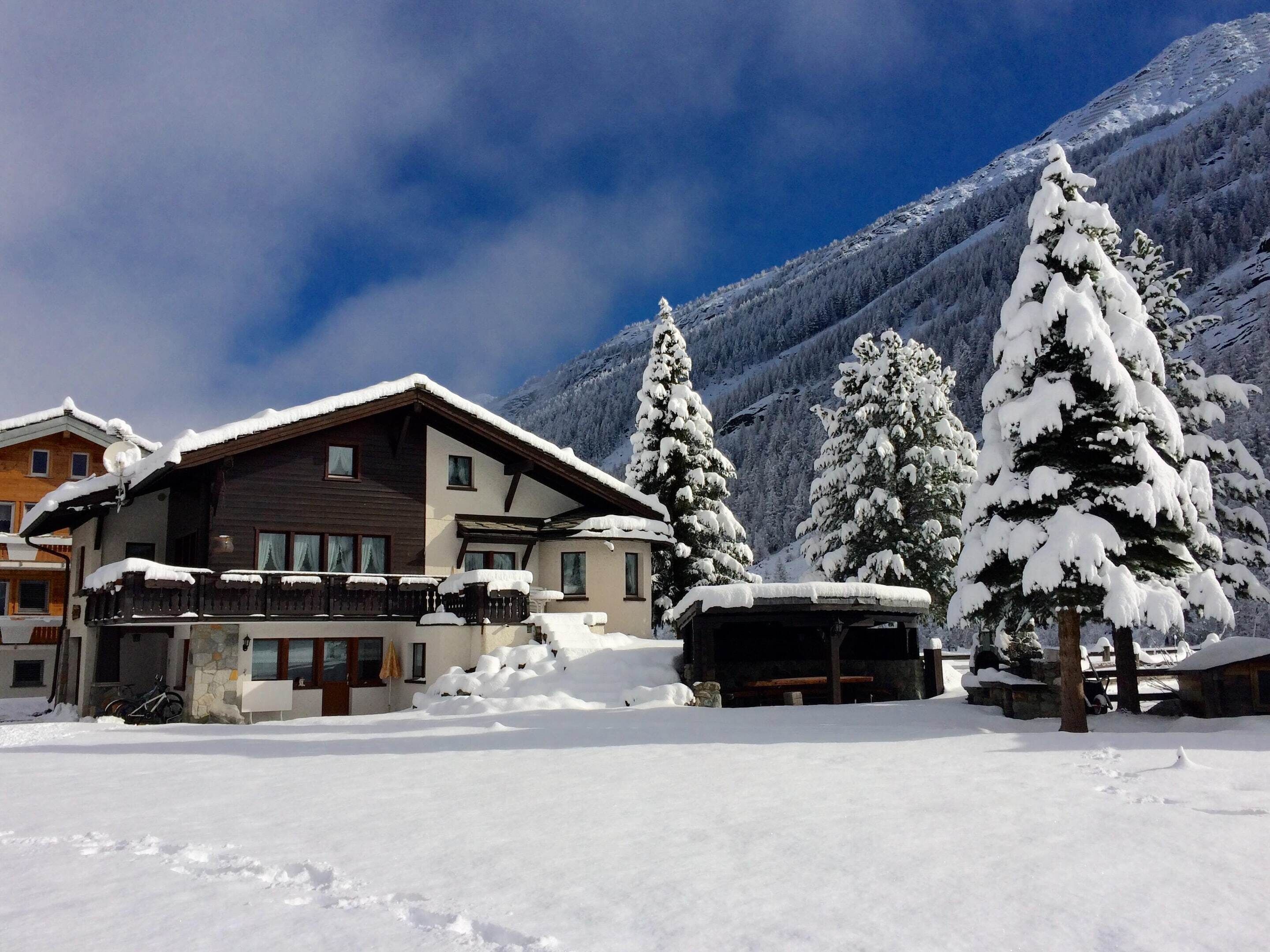 Un edificio cubierto de nieve con un balcón, rodeado de árboles y montañas cargadas de nieve.