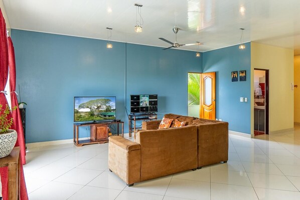 A living room with a blue wall, a brown sofa, a TV, a ceiling fan, and a door with a plant. - Charming 3-bedroom house with AC and WiFi in Quepos (Quepos)