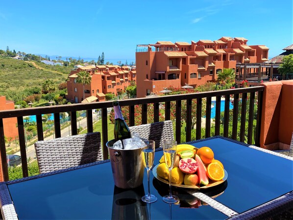 A balcony with a table set for two, a bottle of champagne, and a bowl of fruit. The view includes a pool and orange-colored buildings in the background. - Sea View Penthouse Apartment with Hot Tub near Estepona - Casa Bella Vista (Buenas Noches)