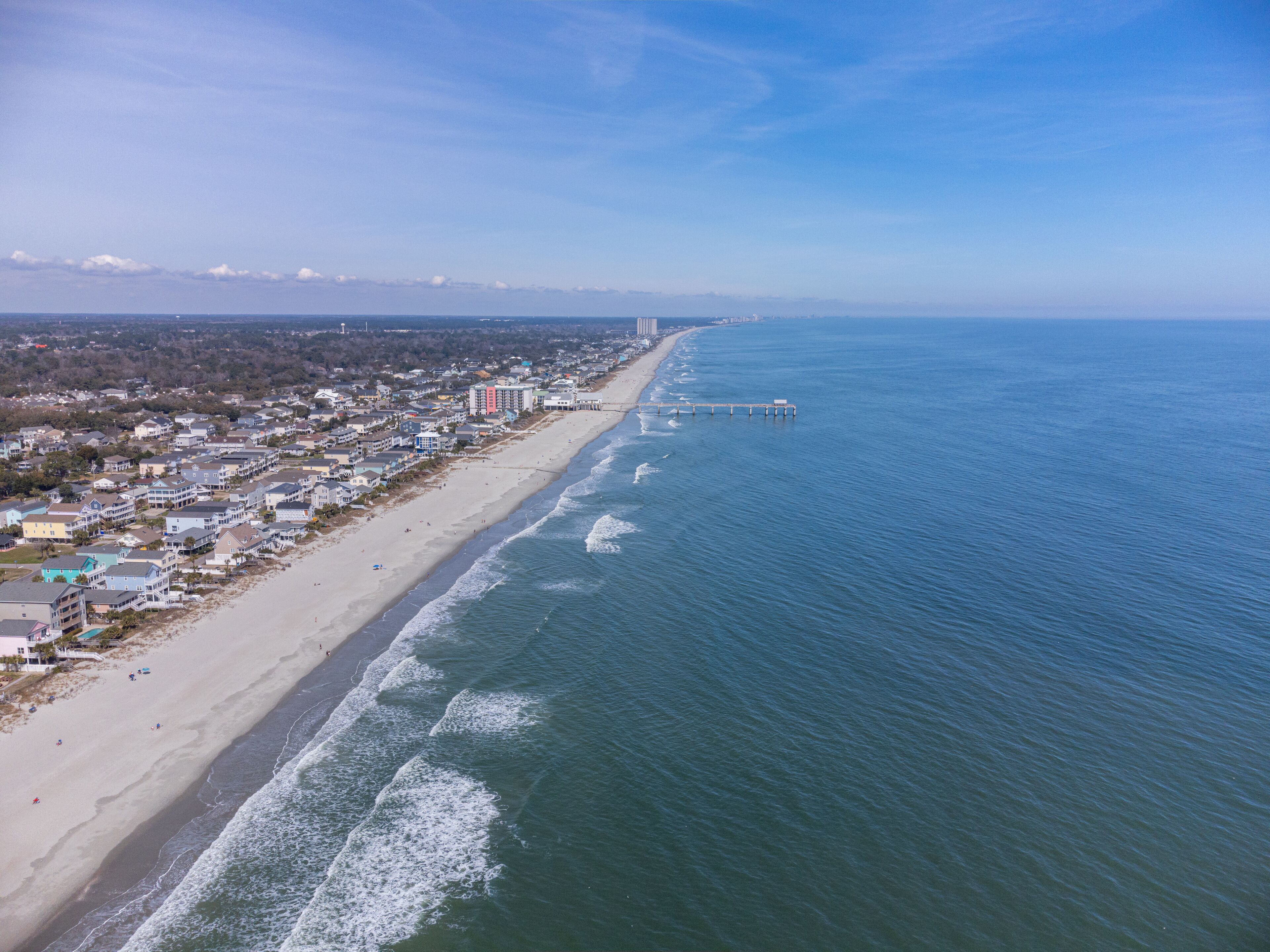 Une vue aérienne d’une ville côtière avec une longue plage, une jetée qui s’avance dans l’océan et des immeubles résidentiels le long du littoral.